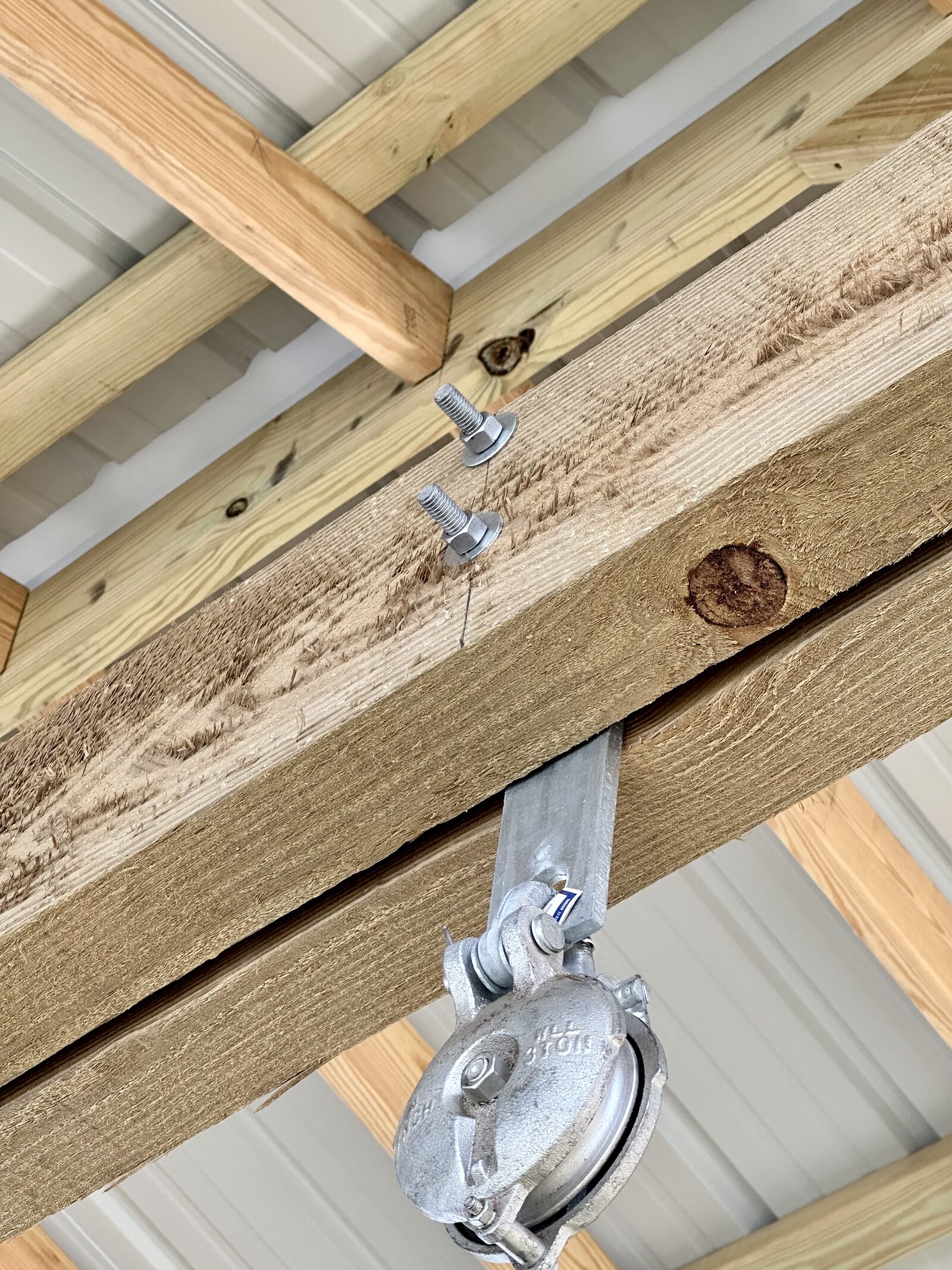 A galvanized boat-lift sheave block hanging from a fabricated galvanized bracket bolted through the timber beam, with two further galvanized bolts in the beam alongside