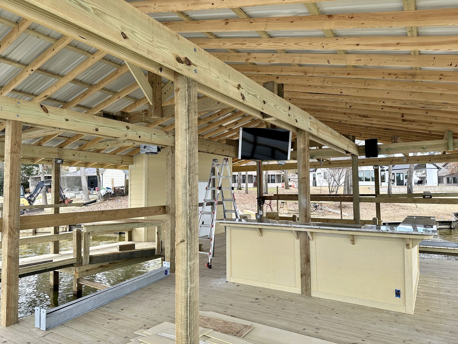 Interior of a boathouse under construction showing the roof framing, timber posts, rails and a kitchen bar, with an excavator working on the bank beyond