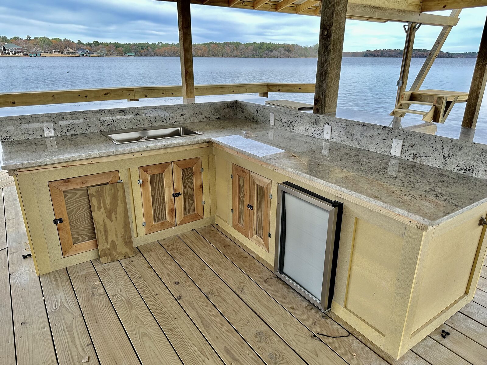 An L-shaped outdoor kitchen on a boathouse deck with granite counters, a stainless sink, an under-counter refrigerator and weather-rated outlets, looking out over the water
