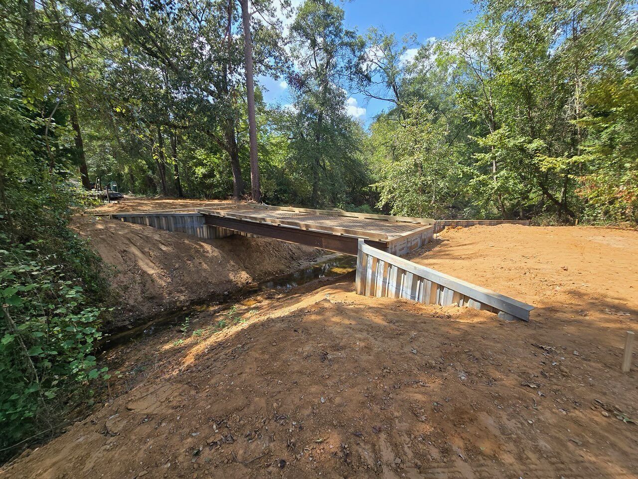 The concrete cap and curbs of a completed steel bridge from the approach, galvanized sheet-pile wingwall on the right and the raw earth approach still to be graded