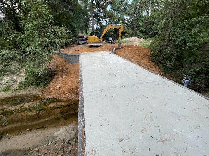 A freshly poured concrete bridge deck curing on its steel girders, sheet-pile wingwalls either side and an excavator at the far end of the span