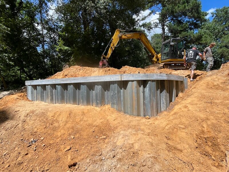 A galvanized steel sheet-pile buttress wall being driven at a bridge abutment, an excavator and crew working from the top of the cut bank