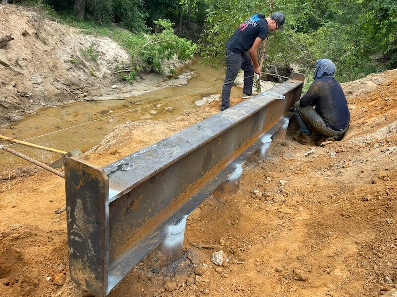 Two crew members welding a steel landing beam to the tops of driven pipe piles at a bridge abutment, the creek bed visible below