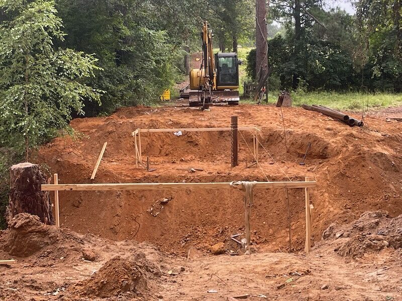 A single steel pipe pile driven at a bridge abutment, held plumb in a timber template with string lines, an excavator working on the red-clay bank behind