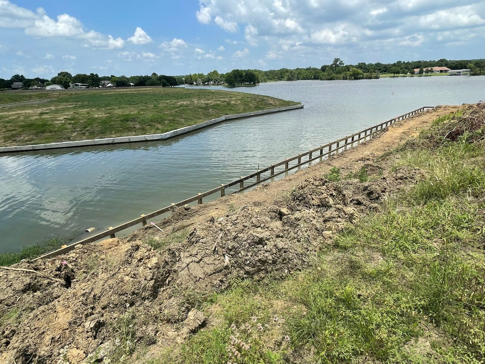 Wide view across a subdivision lake with a concrete-capped bulkhead wrapping the far shoreline and new timber bulkhead framing along the near bank