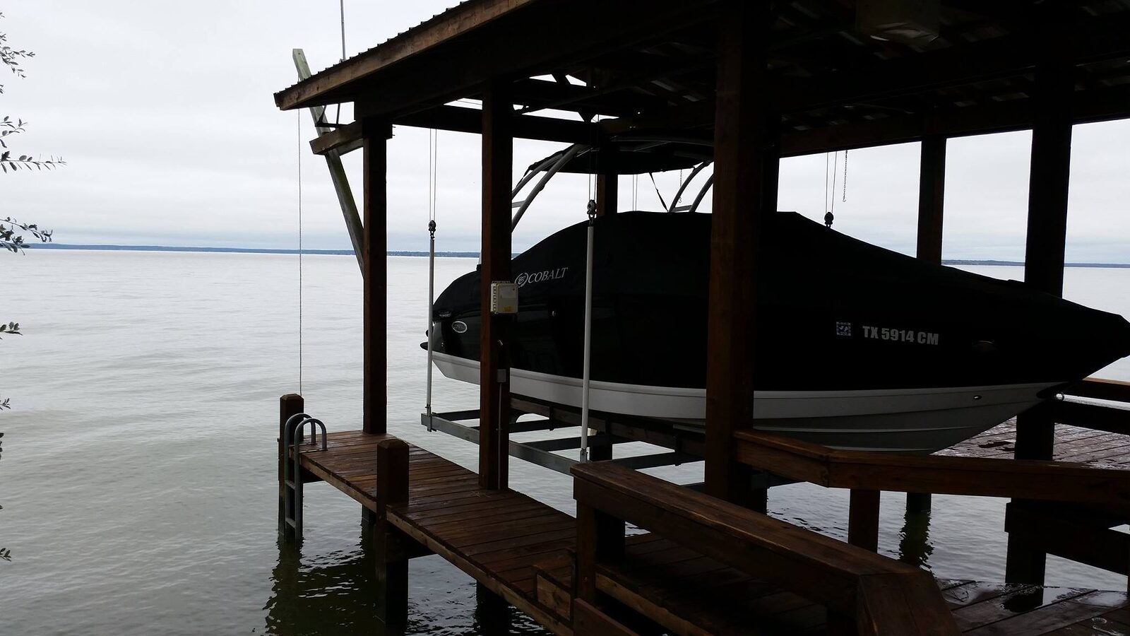 Covered boat rests on a lift inside a timber boathouse slip, with the walkway and pile line running out into open water