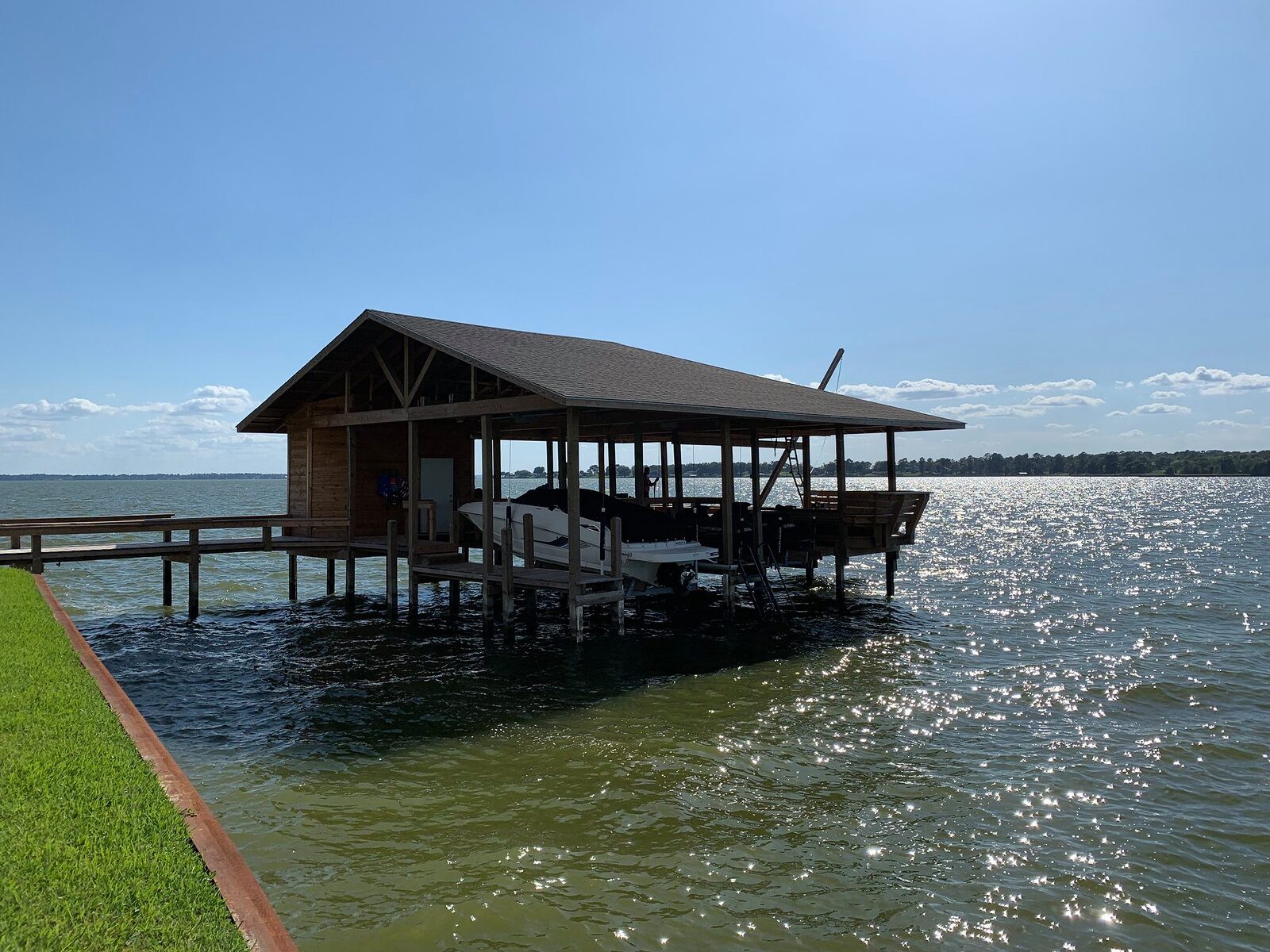 Covered boathouse standing on timber pilings in shallow lake water, reached by a walkway from the lawn
