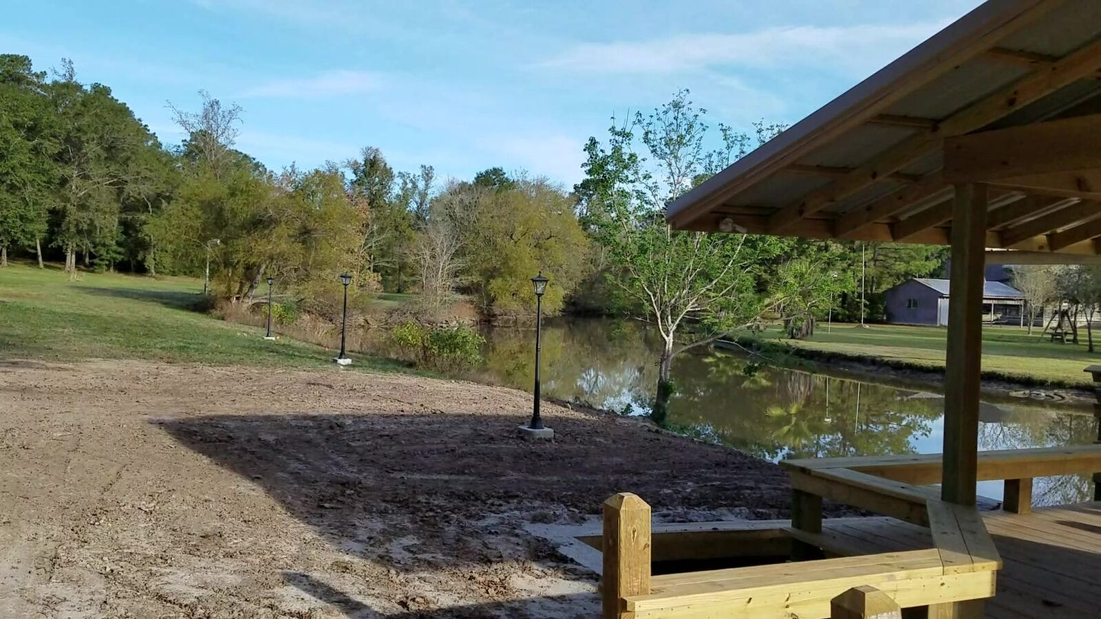 The overhanging roof and new deck of a covered boathouse frame the right side of the shot, looking across still water to a lawn with lamp posts and trees.
