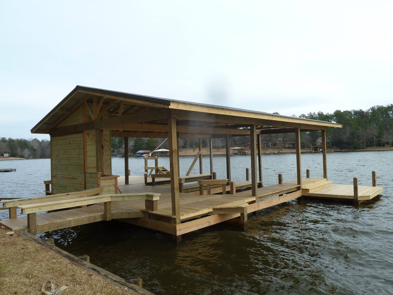 Completed covered boathouse with a wide surrounding deck and a timber walkway ramping down to it from a private lakefront bank