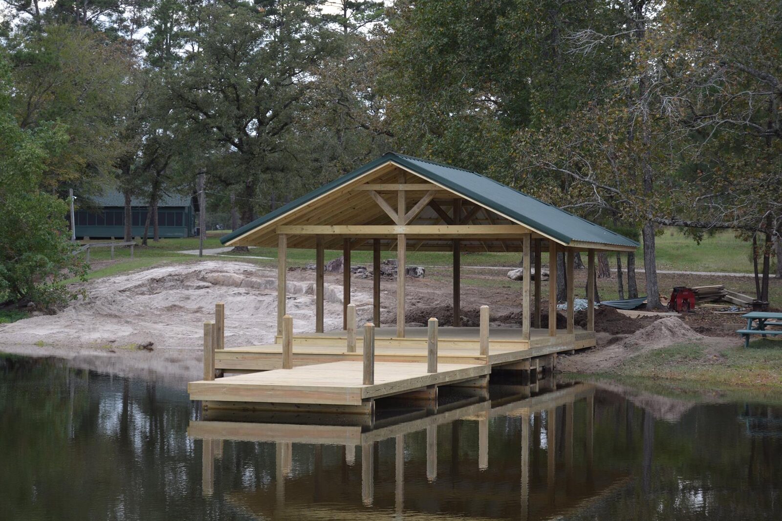 Covered waterfront shelter with a green metal gable roof and attached wooden dock reflected in calm water
