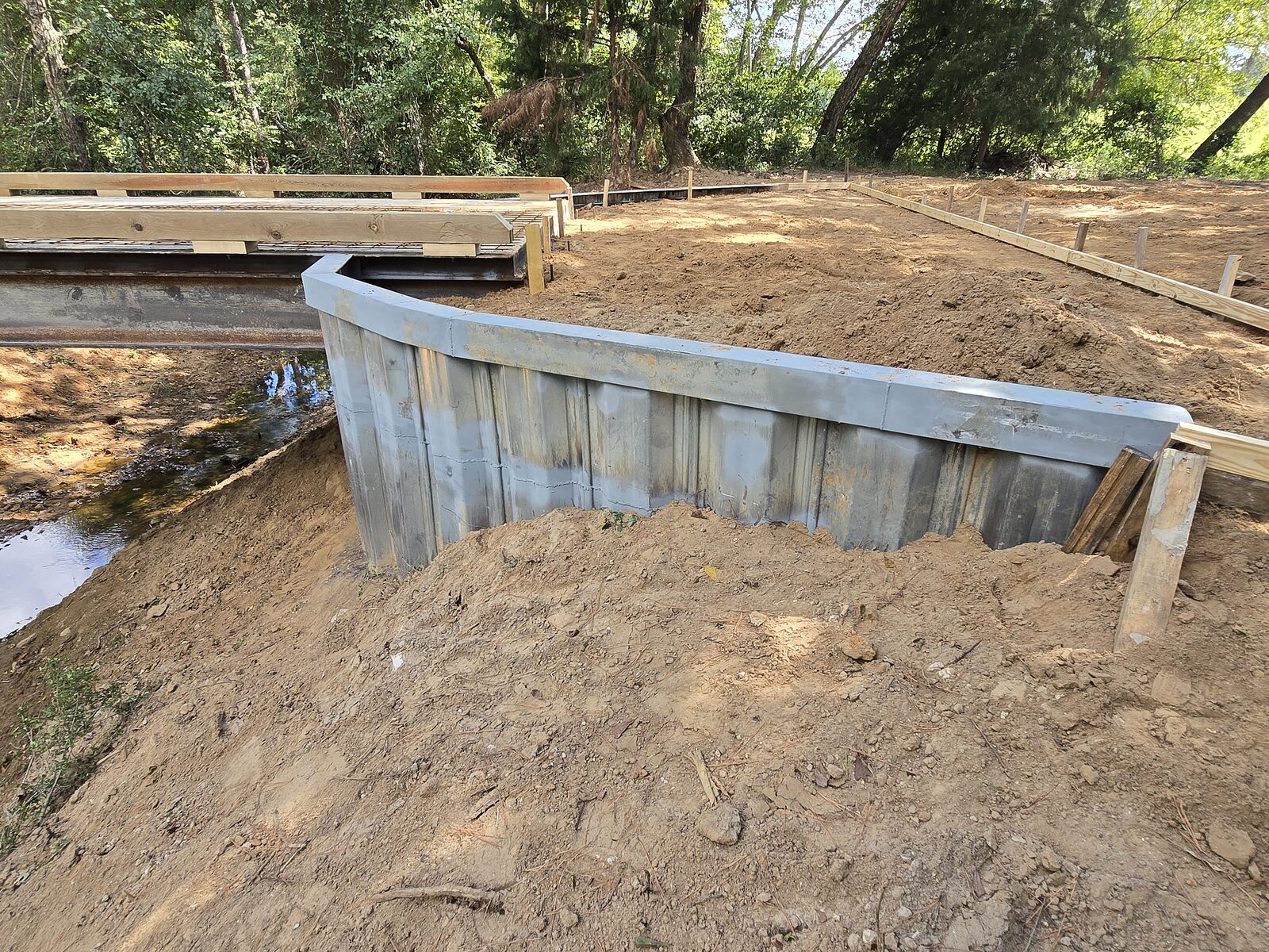 The steel sheet-pile wingwall of a creek crossing seen from the approach, retaining the graded bank where the deck meets solid ground