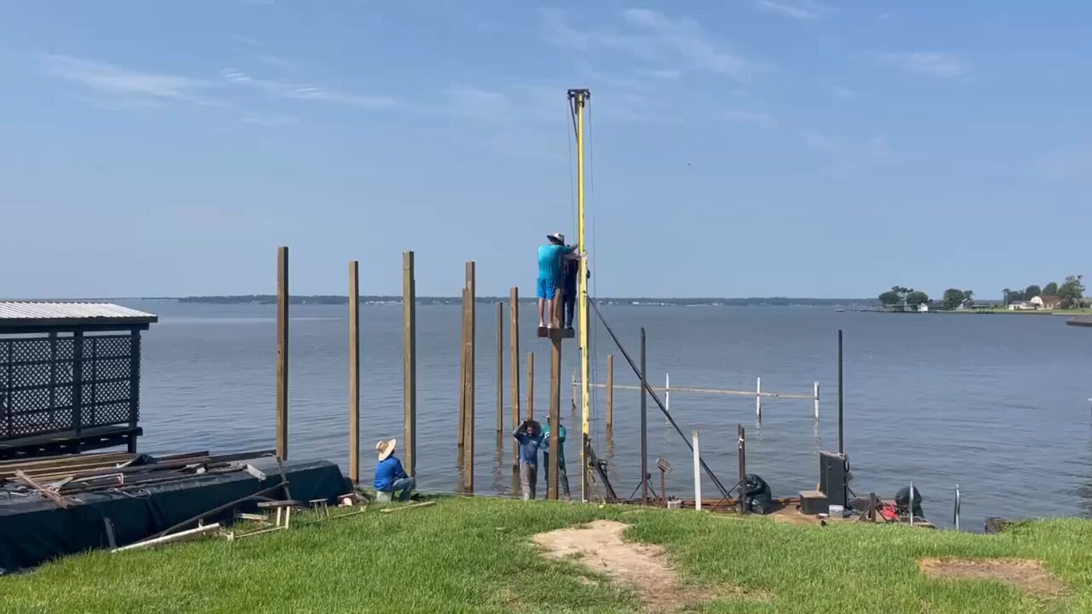 A Shoretech crew driving timber pilings in shallow water with a rig, a line of set piles already standing off the bank