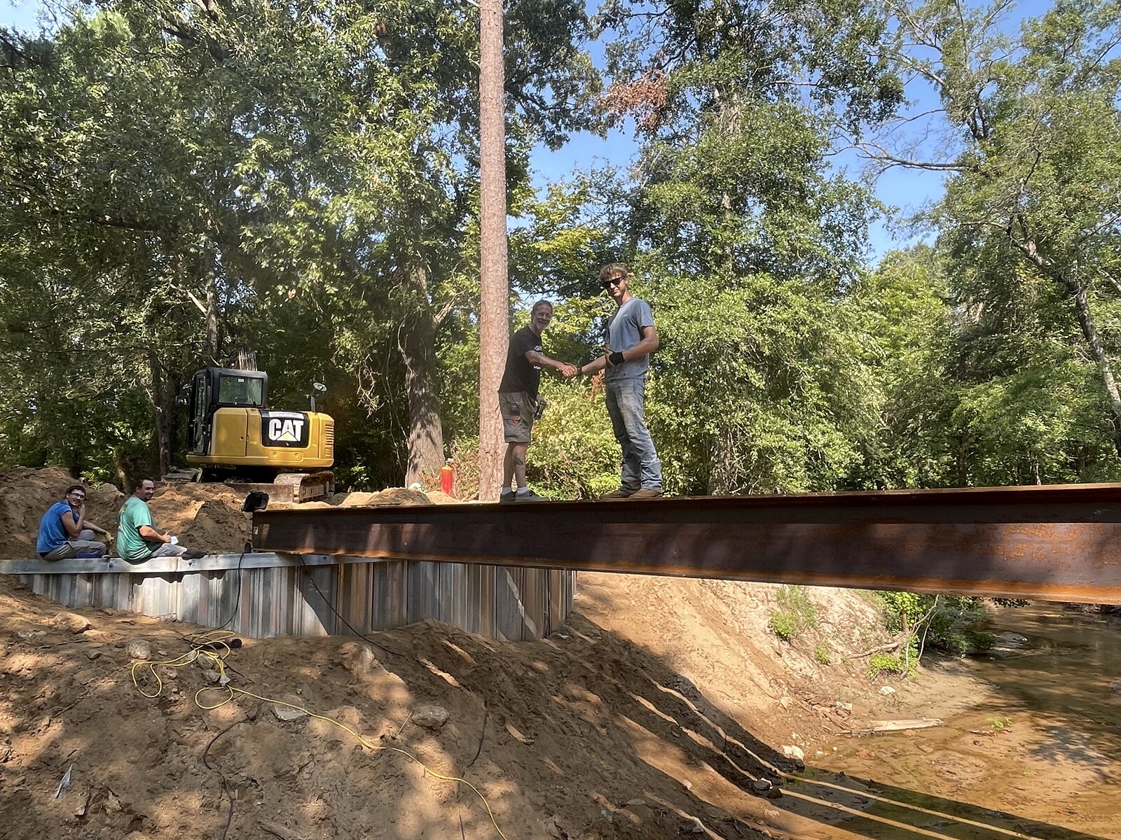 Two Shoretech crew members standing on a freshly landed steel I-beam where it bears on a galvanized sheet-pile buttress wall, an excavator on the bank behind them and the bare red-clay approach cut in the foreground