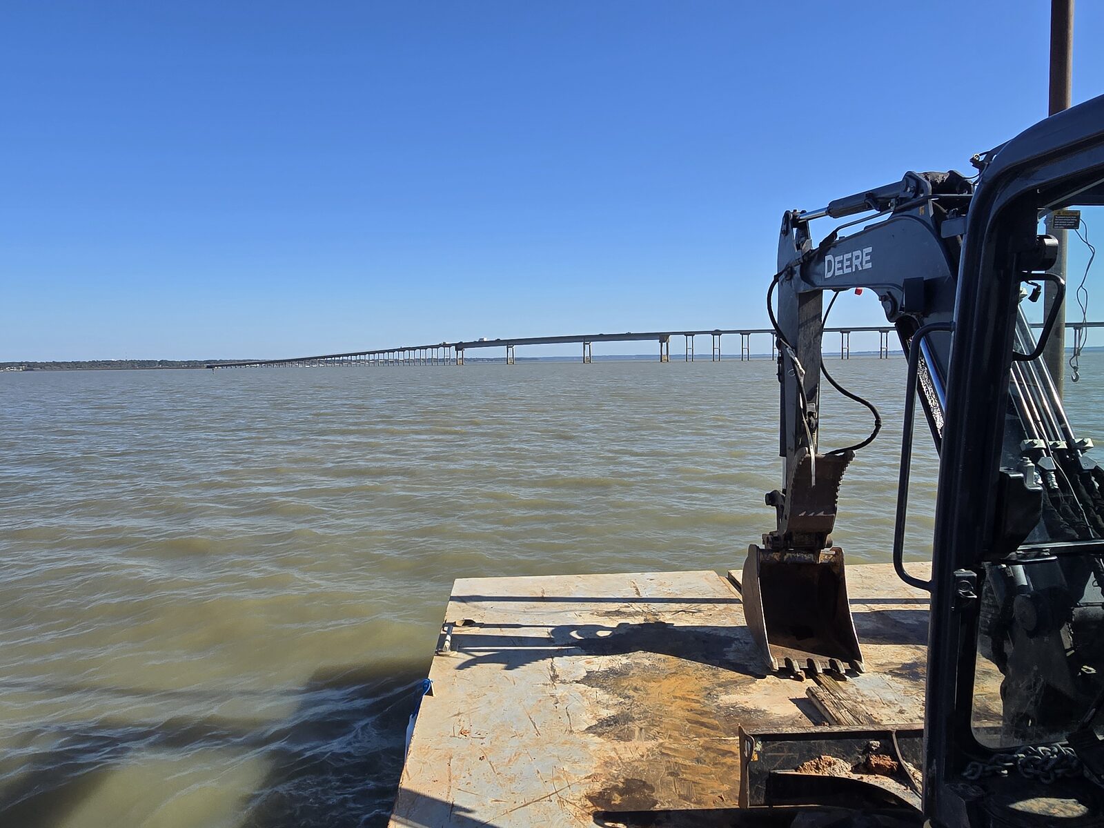 Excavator and bucket on a work barge deck, looking out across open lake water toward a long causeway bridge