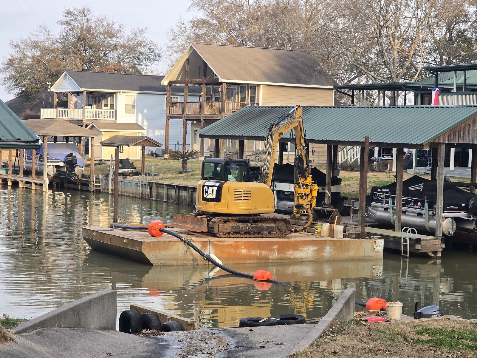 A dredge barge and excavator working the water between two covered residential boathouses, floats holding the discharge line clear of the slips