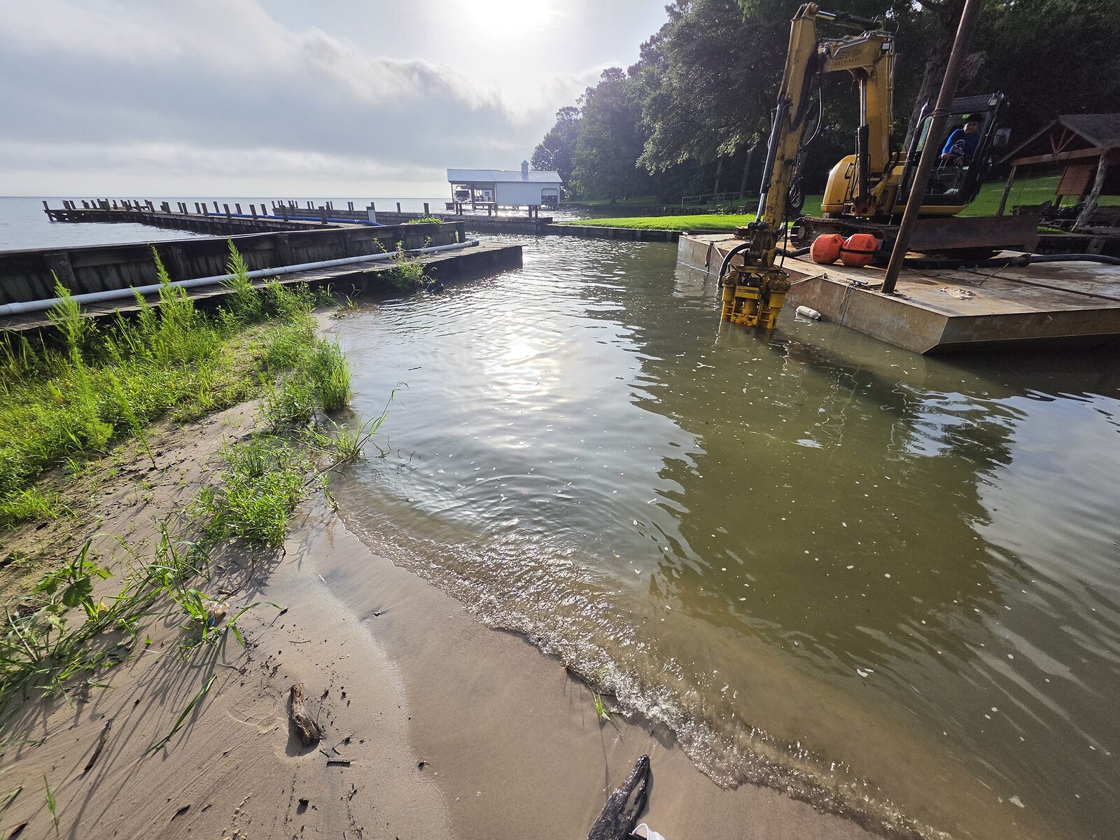 An excavator on a spud barge cutting a shallow slip back to depth beside a residential bulkhead, the pump head down in the water and a pier running out beyond