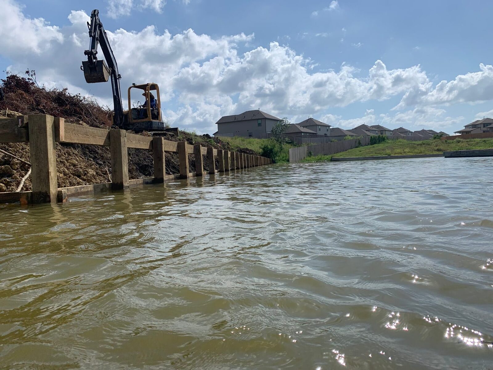 Excavator working on the bank above a long run of newly installed timber bulkhead, seen from the water, with subdivision homes behind