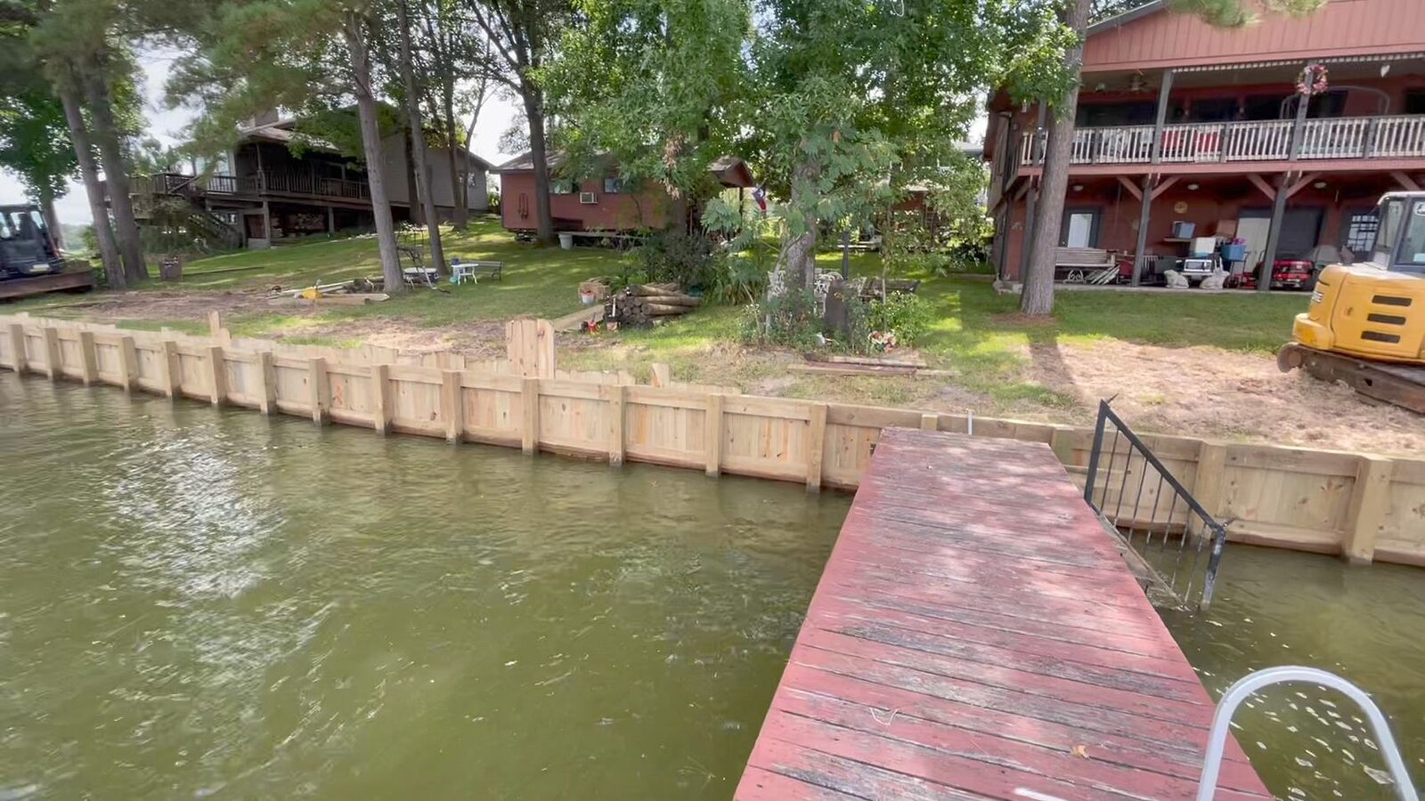 Freshly built light timber bulkhead along a low wooded bank beside an existing red-stained dock, with lake homes on the slope above