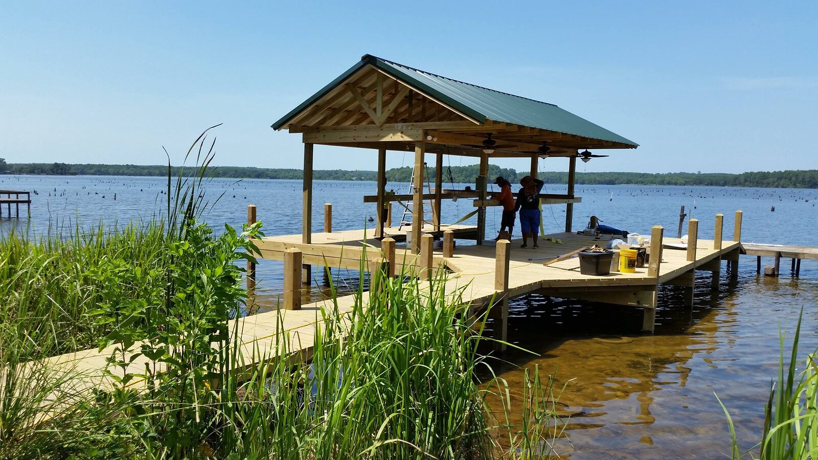 Green metal-roofed timber pier pavilion in use, with a wide plank boardwalk approach crossing marsh grass and an open pier running off to the right