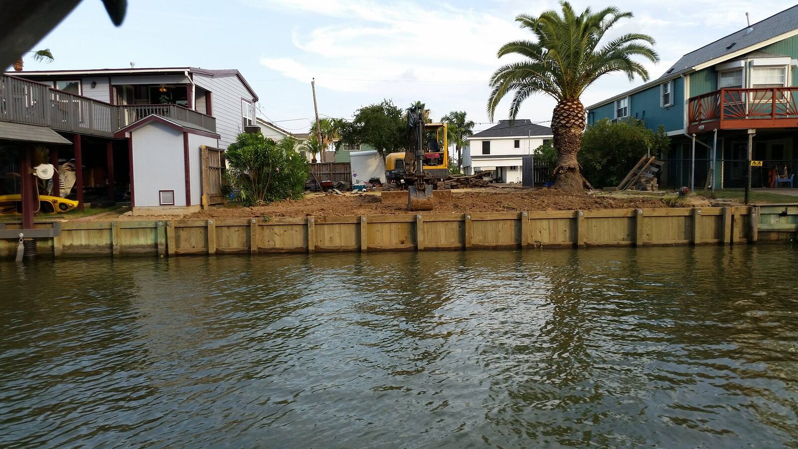 A continuous new timber bulkhead fronting a row of neighboring waterfront houses, seen from the water with an excavator working behind the wall