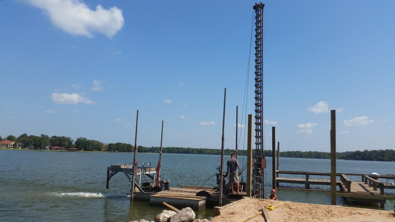 Pile-driving rig on a work barge with tall leads setting piles alongside an existing pier, spray at the waterline under a blue sky