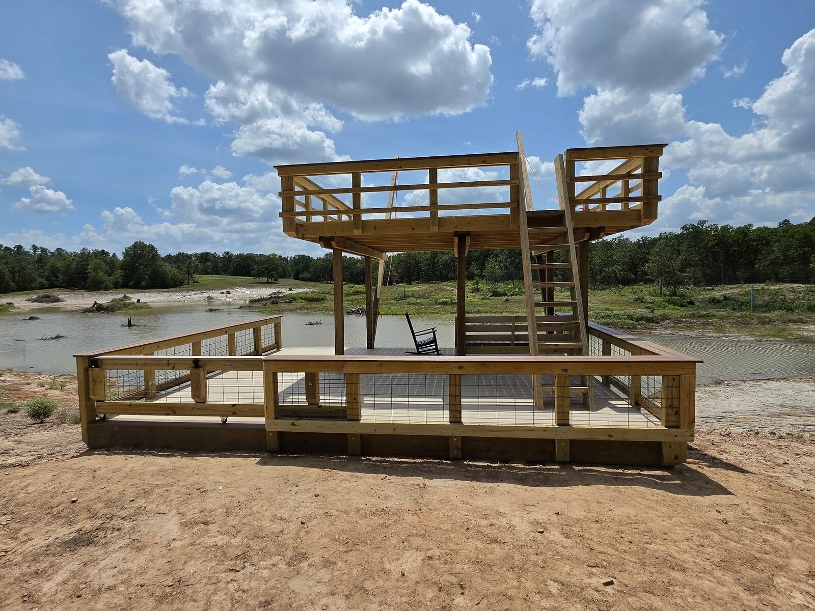 Straight-on view from the bank of the completed two-level private pond swim deck, showing both deck levels, the stair, the ramped side and open water behind
