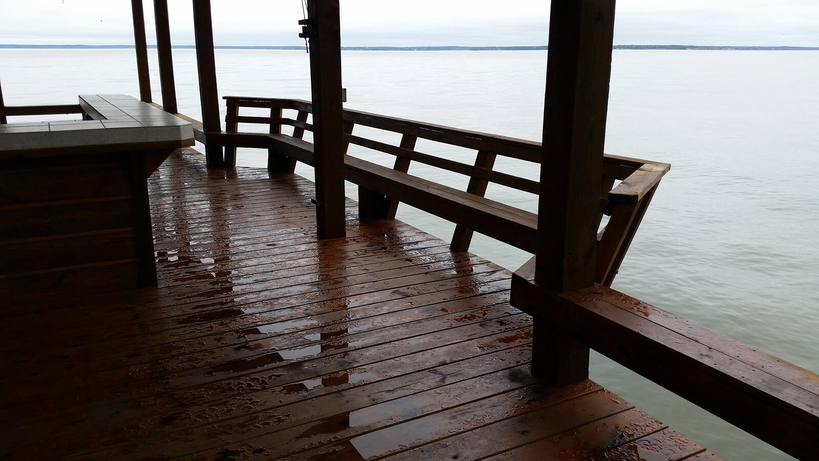 Rain-wet plank decking under a covered structure with a timber bench and horizontal rail, looking out to flat open water under a grey sky