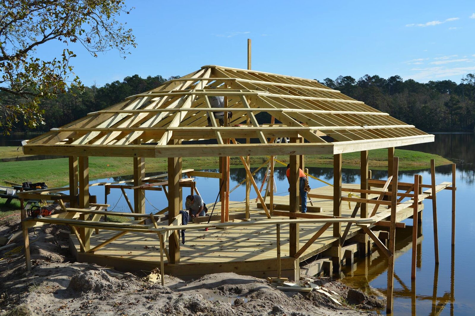 Carpenters framing the hip roof of a waterfront shelter on a completed timber deck above the water