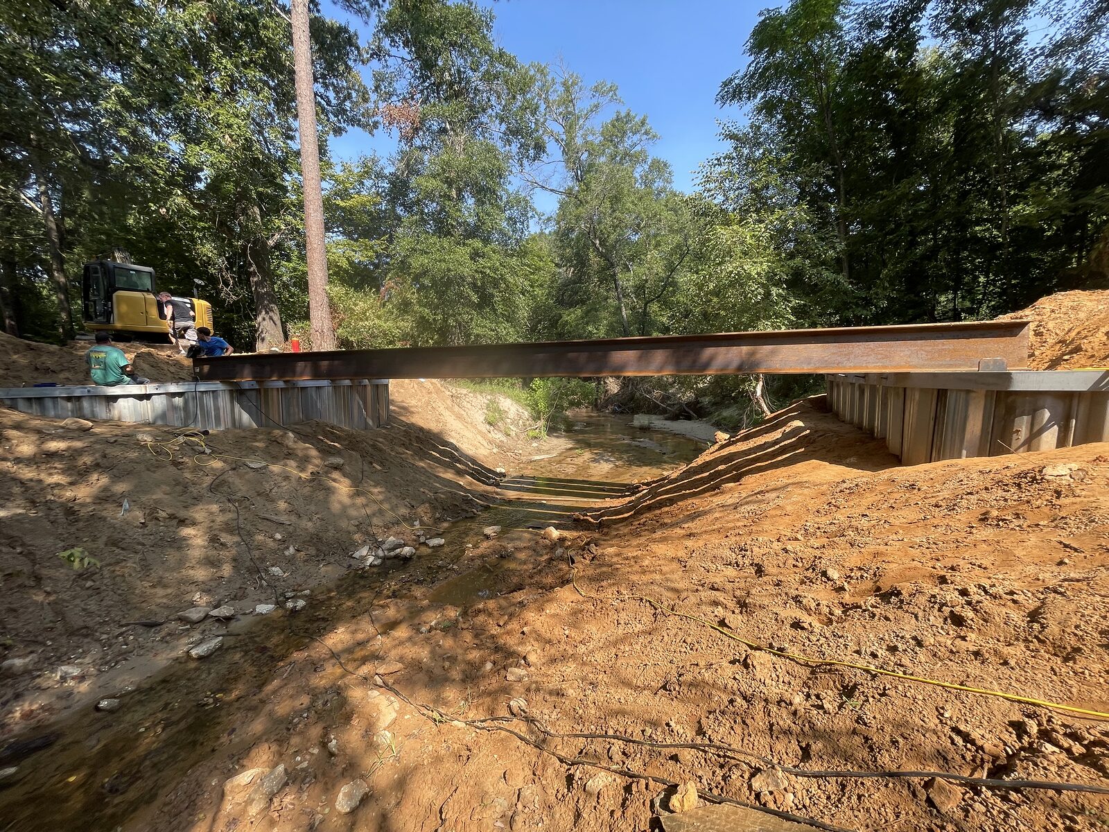 A single steel I-beam girder spanning the full width of a creek from one galvanized sheet-pile buttress wall to the other, seen from the bank below with an excavator at the far abutment