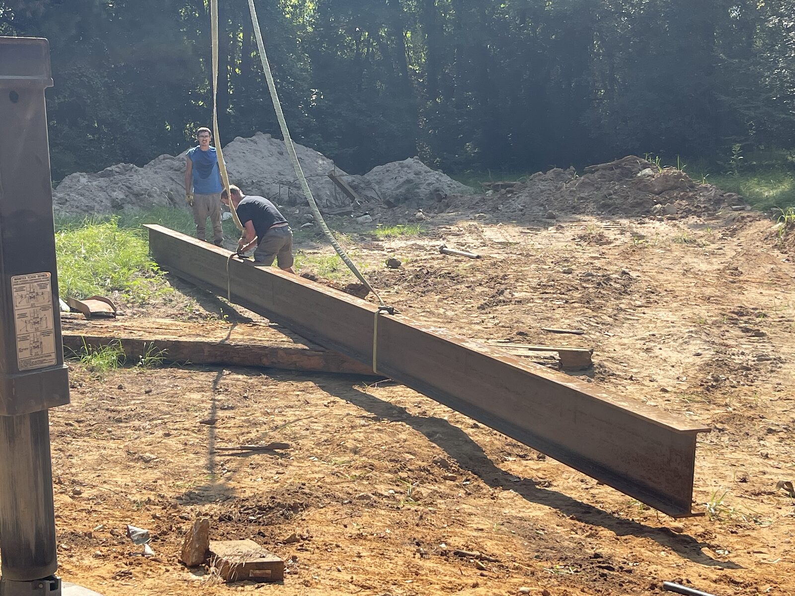 A long steel I-beam suspended from crane straps just above the ground, a crew member steadying one end before it is swung out over the creek