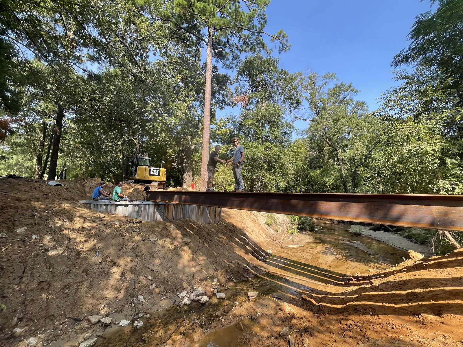 An excavator swinging a steel bridge girder out over the creek toward the sheet-pile abutment while the crew waits to land it
