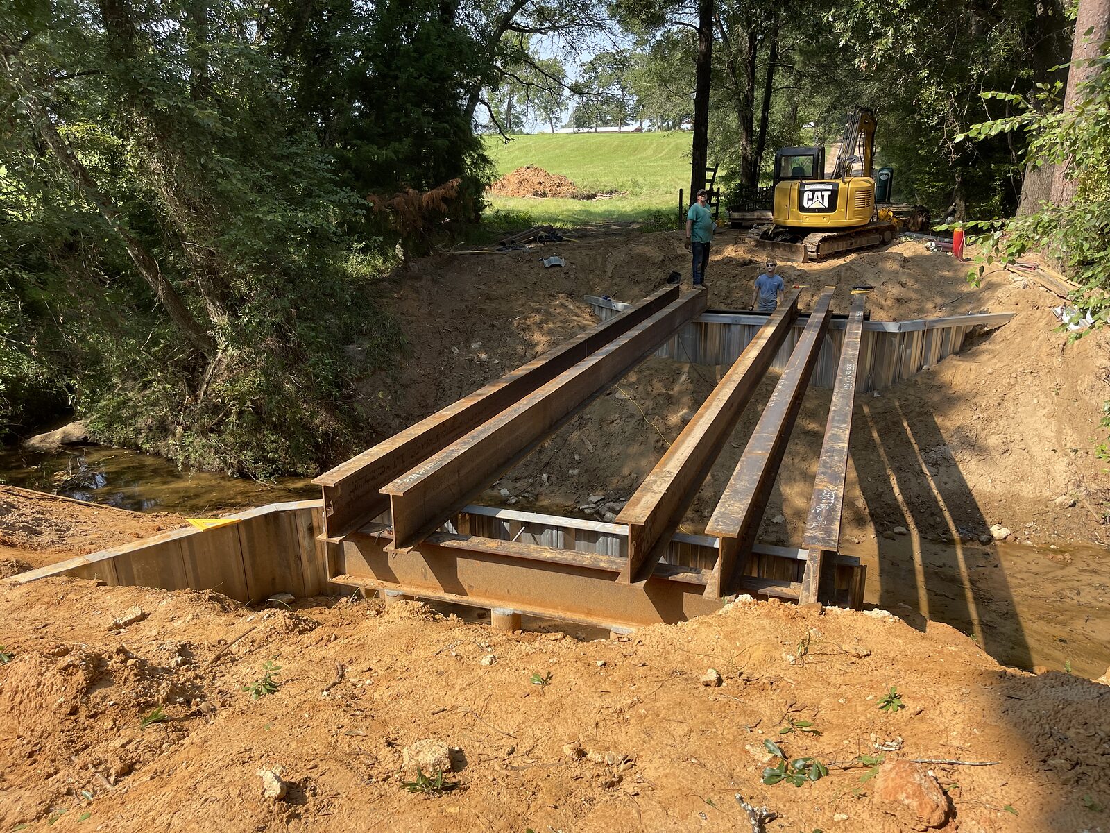 Five steel I-beam girders landed side by side on a galvanized sheet-pile buttress wall, spanning the creek to the far abutment, excavator parked on the bank