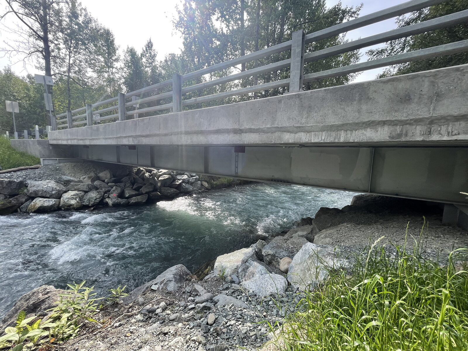 A completed county road bridge with a concrete deck and parapet and steel guardrail on steel girders over a fast, rocky creek