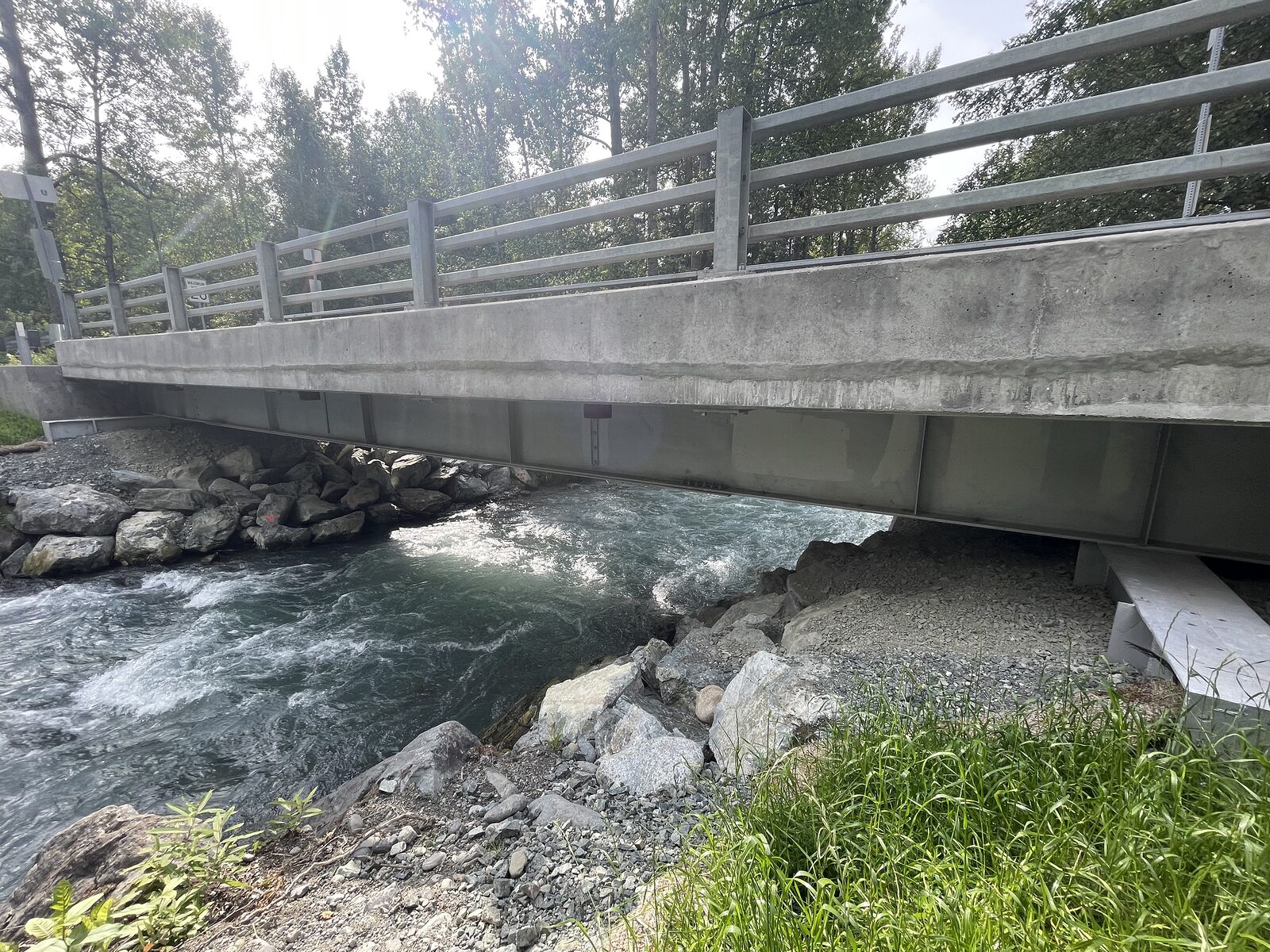 A completed county road bridge with a concrete deck and parapet and a steel guardrail, carried on steel girders over a fast, rocky creek, riprap placed along the bank beneath the abutment