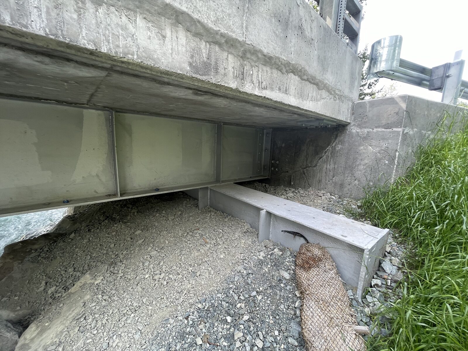 Looking up at the underside of a finished bridge: steel I-girders bearing on a concrete abutment beneath the concrete deck, gravel and drainage fabric at the bank
