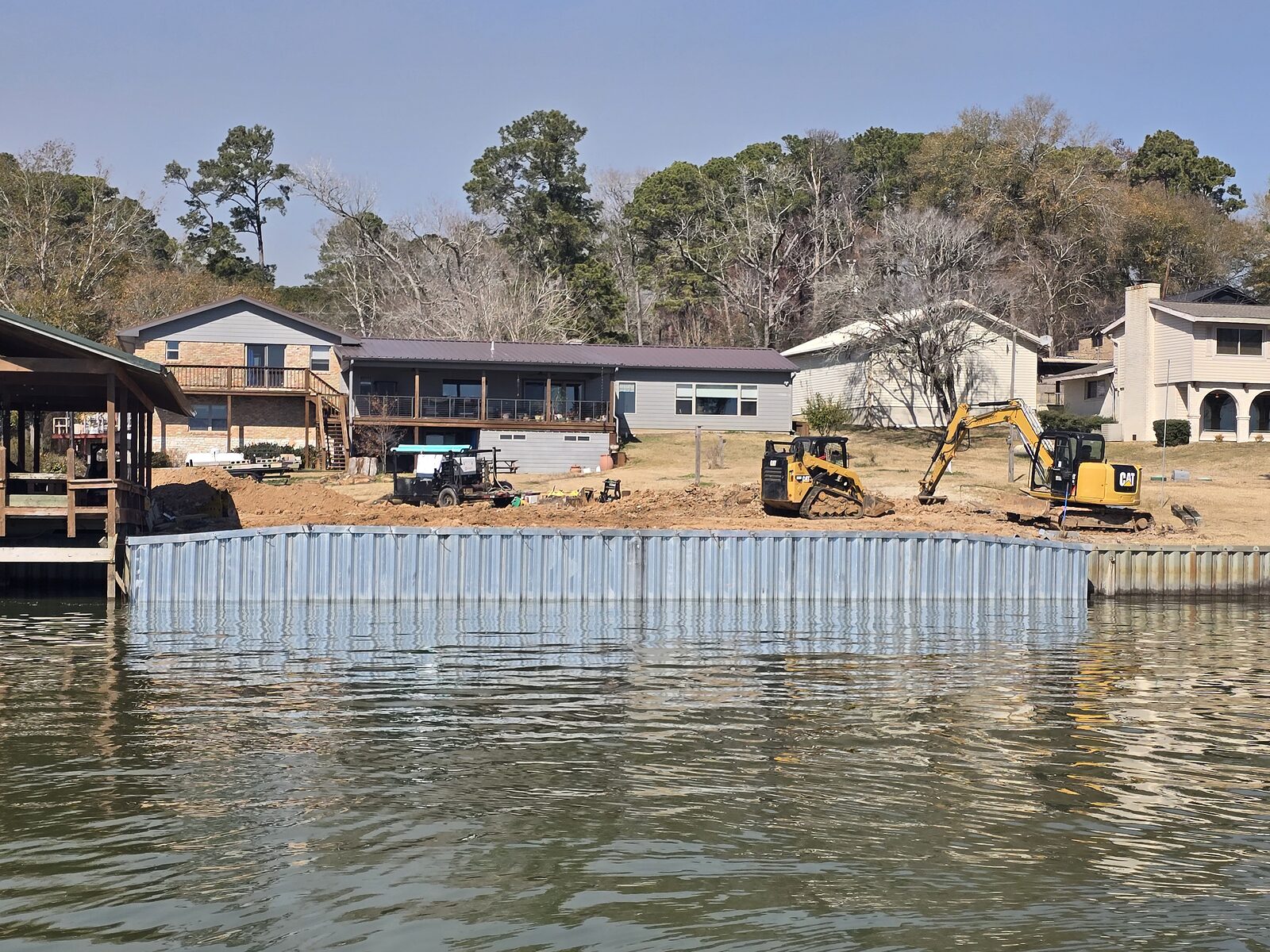 New steel bulkhead running along a subdivision waterfront, with an excavator and skid steer grading the backfill behind it and houses beyond
