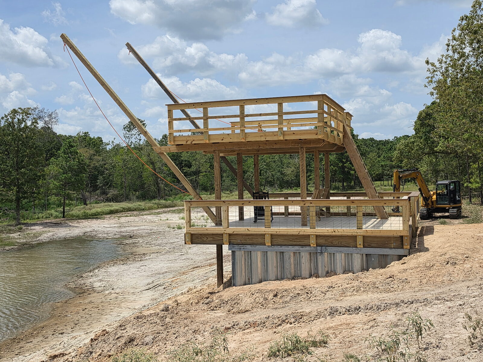 Side view of a two-level private pond swim deck whose entire base is a driven corrugated galvanized sheet-pile cell, with timber swing arms rigged above the deck