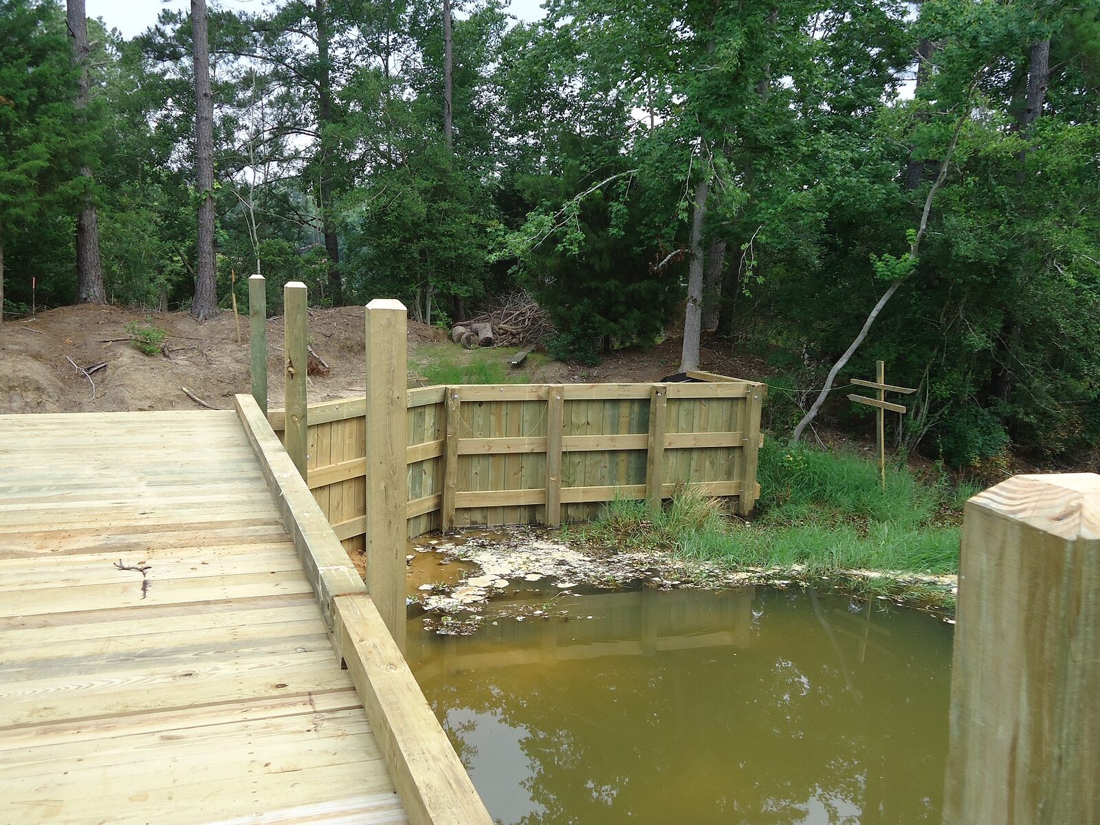 The approach end of a timber crossing where the deck meets the bank, with a timber wingwall retaining the fill either side of the abutment