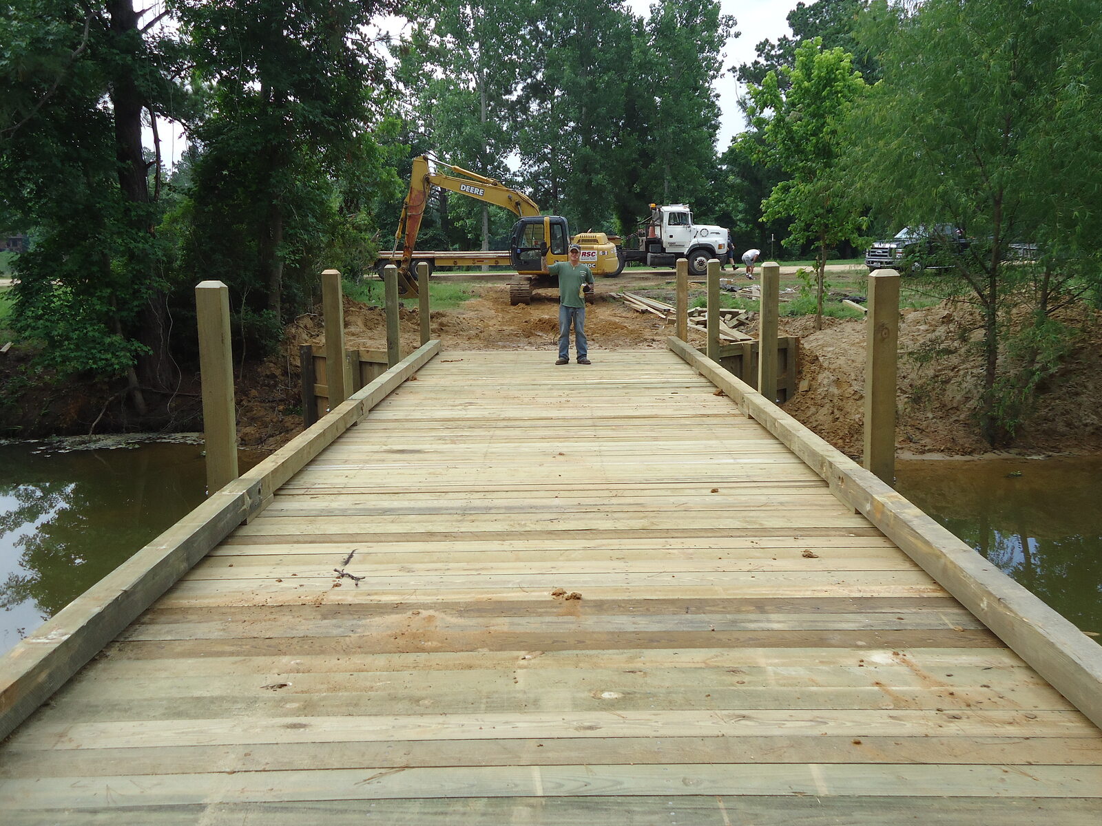 A new heavy-timber bridge deck with square post-and-rail sides seen from the approach in Montgomery County, Texas, a crew member standing at the far end with an excavator and flatbed truck behind