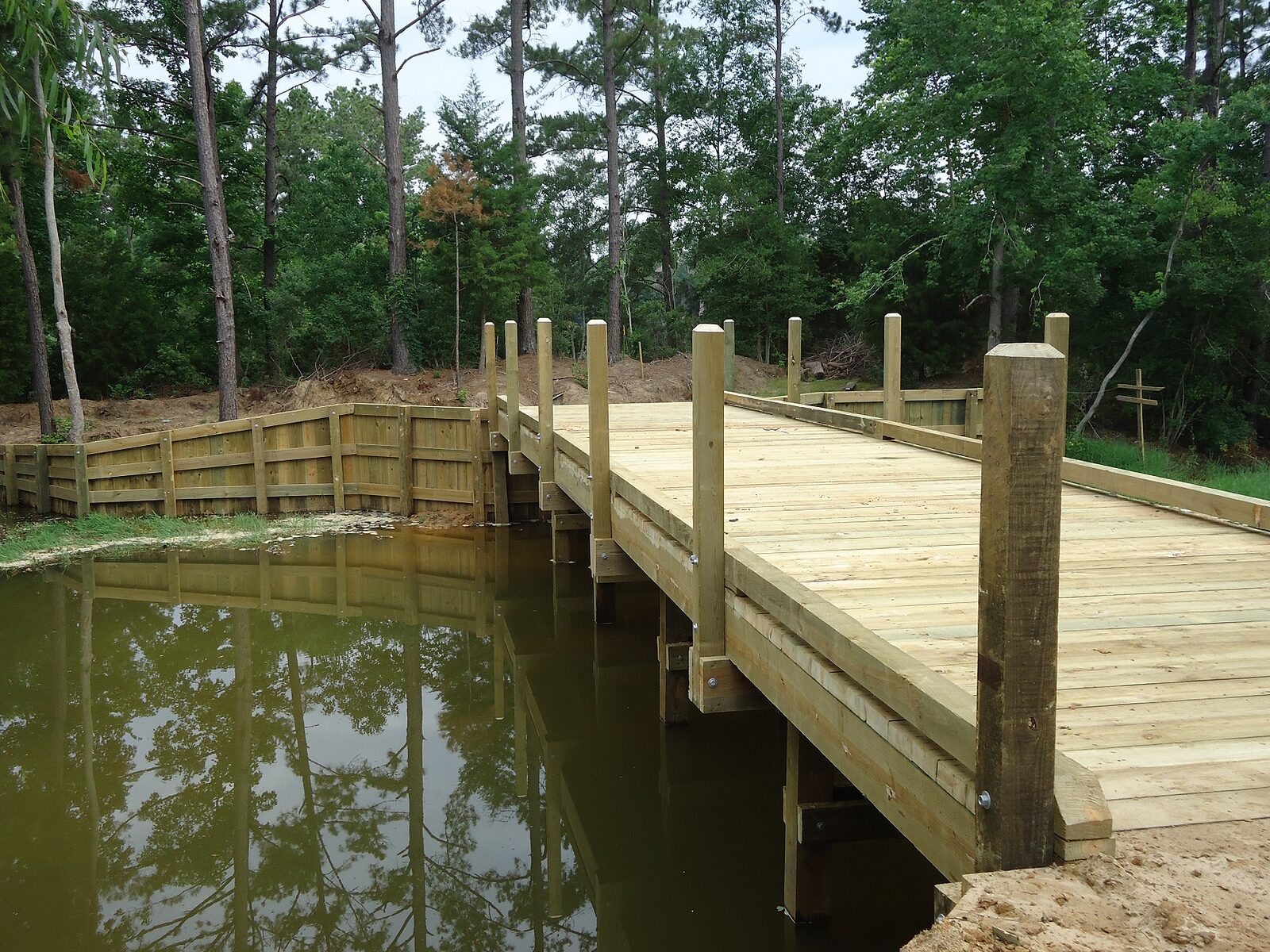 A new timber vehicle crossing under construction, wide deck planking laid over the stringers and rail posts standing, with the retained bank running away on the left