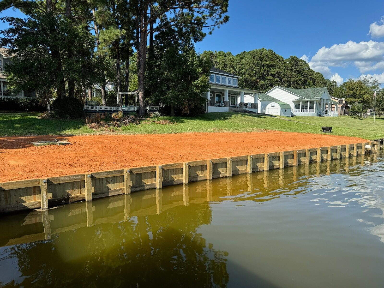 New timber bulkhead running the length of a lakefront lawn with evenly spaced tieback posts along the face and fresh sand backfill graded flat behind it