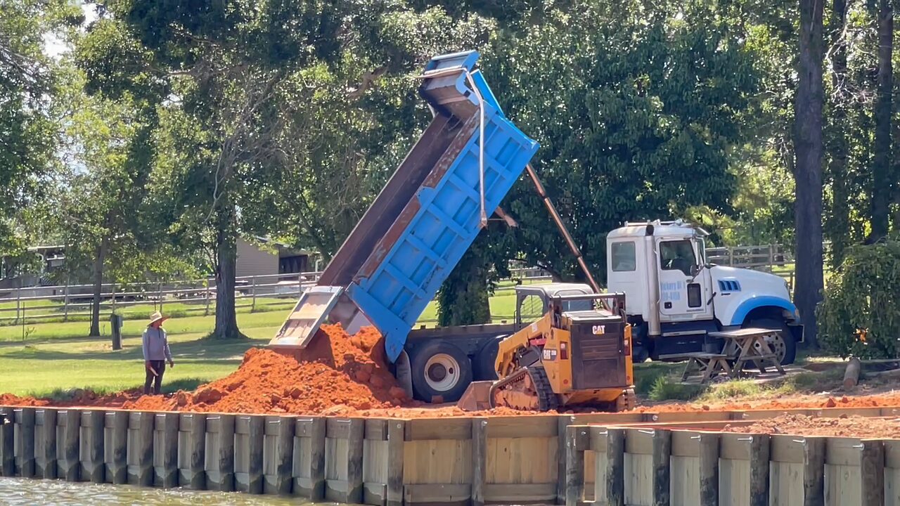 A dump truck tipping a load of red clay onto the shore behind a completed timber bulkhead, a dozer spreading the fill along the wall and the lake in the foreground