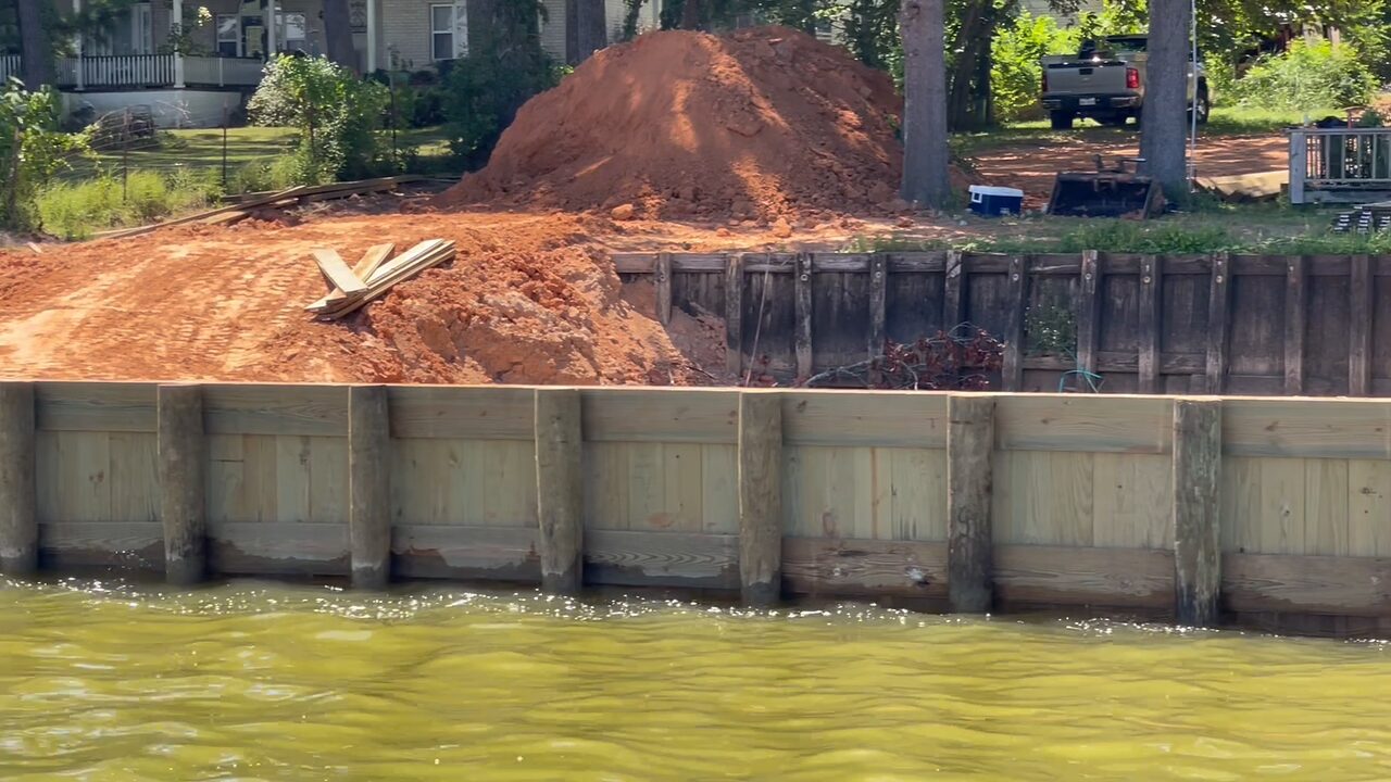 A new heavy timber bulkhead standing in the water with the bank excavated behind it: tieback rods and timber braces still exposed above the wales, a red clay spoil pile and an excavator on the shore