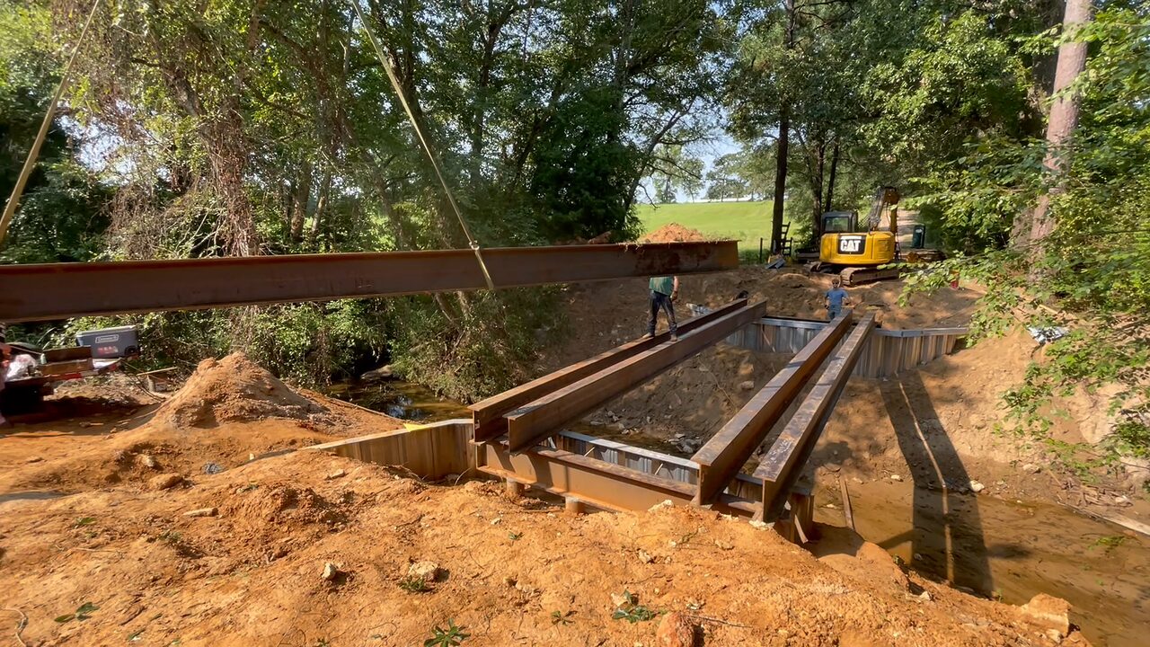 A steel I-beam bridge girder hanging from the excavator's rigging strap, swinging out over the creek toward four girders already landed on the galvanized sheet-pile buttress wall